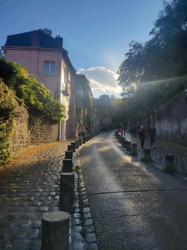 Old cobblestone street with a pink building and trees curving up a hill with susn peeking out from clouds in Montmartre, Paris.