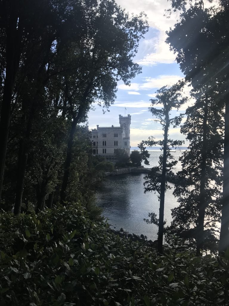 View of a the castle Miramare in Trieste, which is white, with a tower on the shore of the sea, looking through a space in trees with a view of a blue sky and white fluffy clouds.
