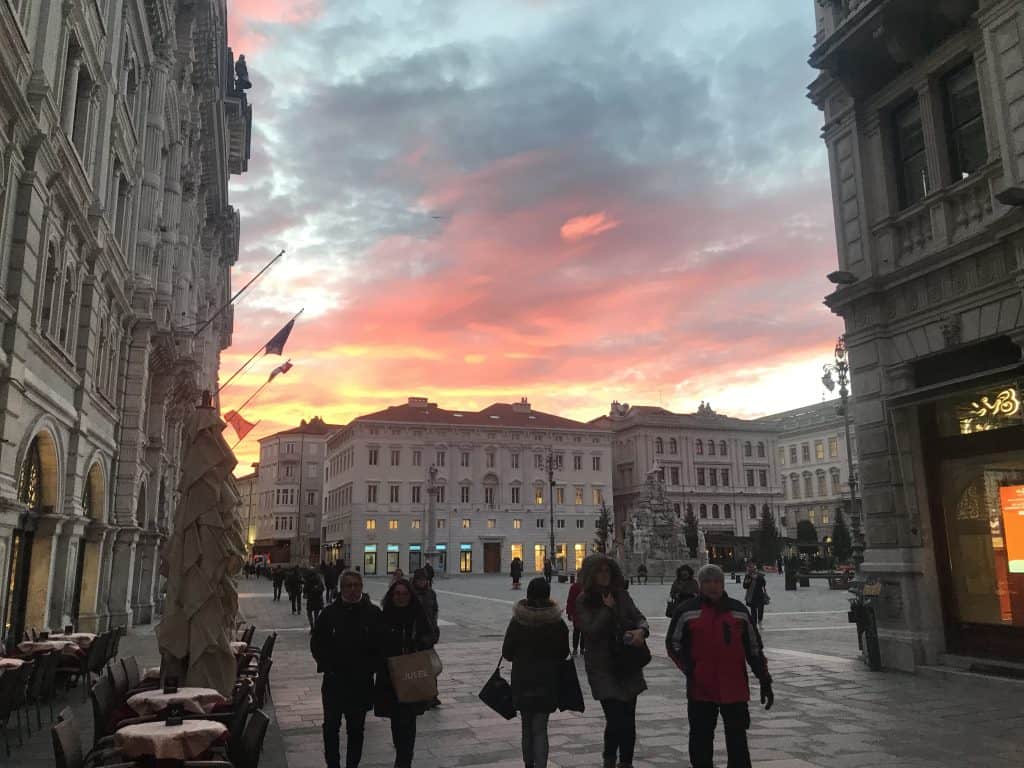 A large plaza in Triest with people walking with coats on, looking out onto baroque buildings and a sunset sky with yellow, pings, oranges and blues.