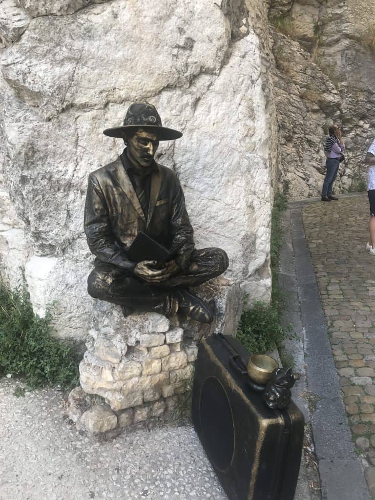 Living statue, a man in bronze clothing and hat with bronze suitcase sitting immobile on stone wall near Pope's Palance in Avignon